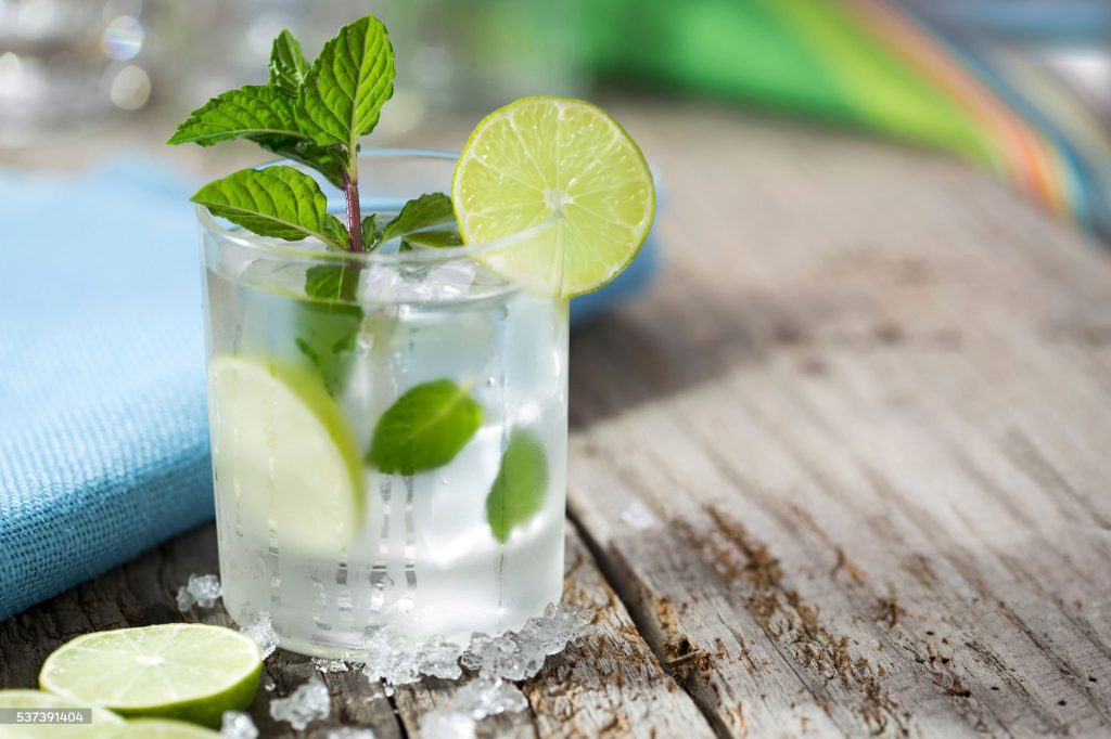 A mojito at the beach with lime and fresh mint with crushed ice on a rustic wood table with the ocean in the background.